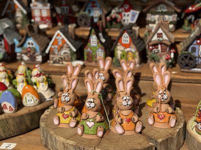 Clay bunnies with painted hearts are displayed on wooden slabs, surrounded by miniature colorful houses in a rustic setting.