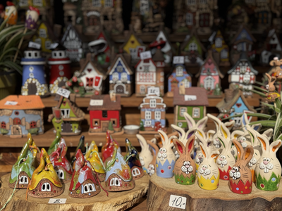 Colorful ceramic figures and miniature houses displayed on wooden shelves at a market stall, each with a visible price tag.