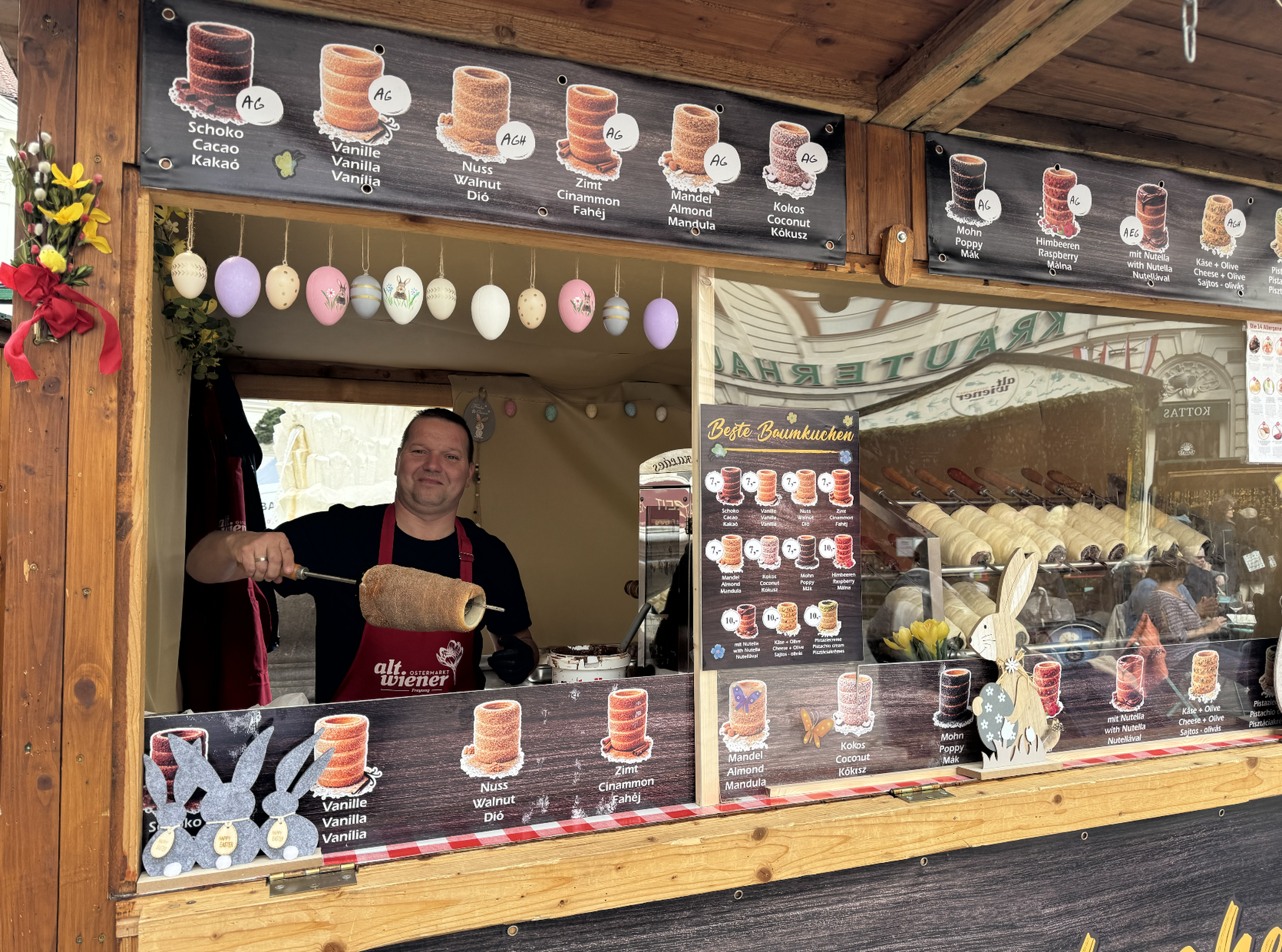 Vendor making chimney cakes at a wooden stall, decorated with colorful Easter eggs and displaying various cake flavors on signs.