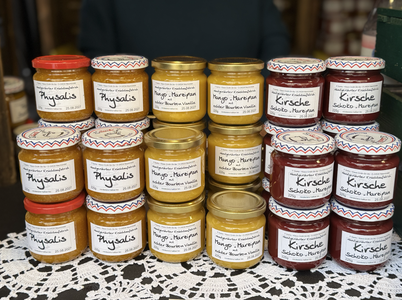 Jars of fruit preserves labeled "Physalis," "Mango-Maracuja," and "Kirsche" stacked on a lace-covered table at a market.