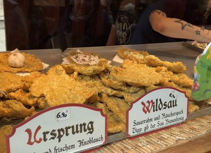 Various fried potato pancakes displayed in a glass case with signs labeled "Ursprung" and "Wildsau." A person is visible in the background.