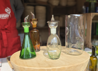 Four glass oil dispensers in various shapes and colors are displayed on a wooden stand in a market.