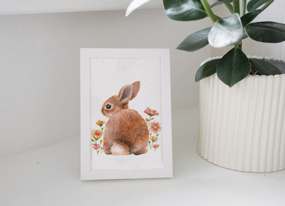 A framed illustration of a brown rabbit surrounded by small flowers, placed next to a plant in a white pot on a shelf.