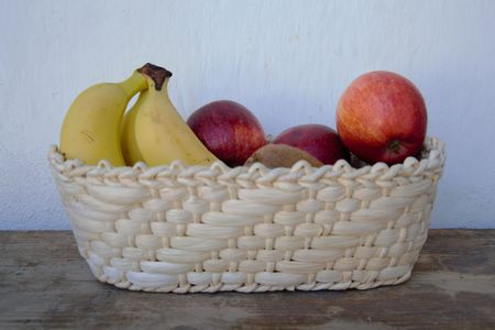 Woven basket with bananas, red apples, and a kiwi on a wooden surface against a white wall.