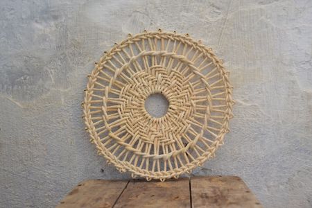 A round, intricately woven natural fiber wall hanging with a central opening, displayed on a textured, light gray wall above a rustic wooden surface.
