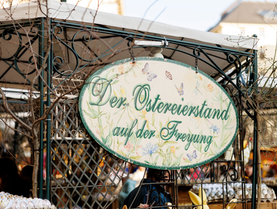 A decorative sign reading "Der Ostereierstand auf der Freyung" hangs at an outdoor Easter market stall, surrounded by branches and people.
