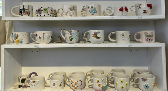 Shelves displaying various handmade ceramic mugs, each uniquely decorated with colorful designs like flowers, animals, and geometric patterns.