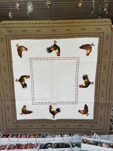 Decorative tablecloth with rooster patterns around the edges, displayed above a shelf with various small items.
