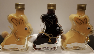 Three bunny-shaped bottles with gold caps, googly eyes, and small bells, containing yellow and dark liquid, displayed on a neutral background.
