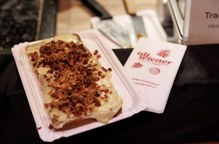 A sandwich topped with melted cheese, crispy bacon, and fried onions on a paper plate, next to a napkin with a red logo.