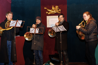 Four musicians play French horns on stage, standing in a row with sheet music, under a decorative cherub.