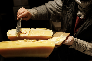 A person scrapes melted cheese from a large block onto a piece of bread, wearing winter clothing and using a spatula.