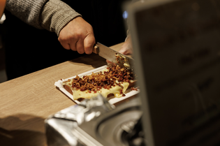 Person slicing a cheese and chopped bacon dish on a wooden countertop, with focus on hands and knife.