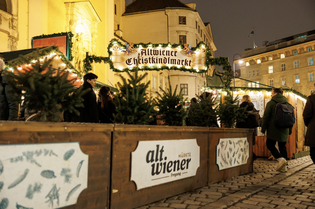 Festive Christmas market scene with illuminated "Altwiener Christkindlmarkt" sign, stalls, people, and decorations on a cobblestone street at night.