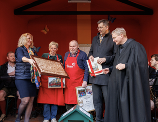 A group of people stands on a stage, holding awards and a carved wooden plaque, with bright red and orange backgrounds.