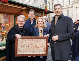 Four people stand smiling outdoors, with one holding a carved wooden artwork of "The Last Supper" in front of a festive market stall.