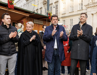 A group of people, including a man in a robe, are standing outdoors near wooden stalls, clapping at an event in an urban setting.