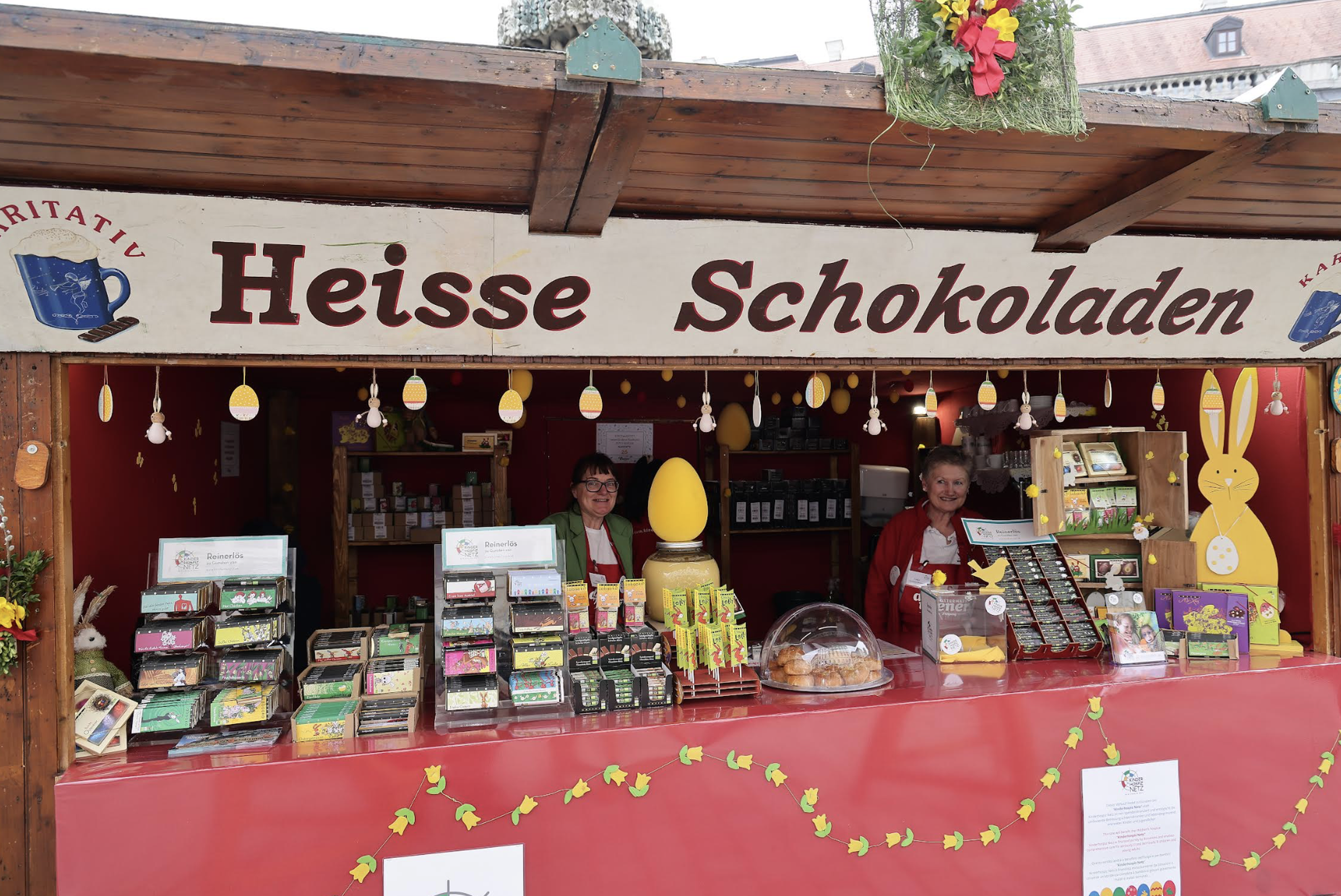 A festive market stall selling hot chocolate, sweets, and Easter-themed items, with two attendants and colorful decorations.