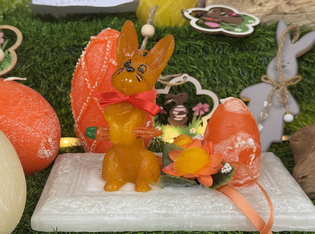 A vibrant orange bunny-shaped candle with a red bow sits on a white platform next to a decorative orange egg with floral accents, set against a grassy background.