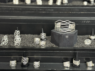 A display of intricate silver rings and bracelets arranged on black stands with a textured background of dark pebbles.