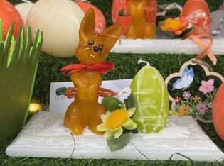 A caramel-colored bunny candle figurine with a red bow holds a carrot next to a green, egg-shaped candy on grass. Nearby, a yellow flower adds brightness.