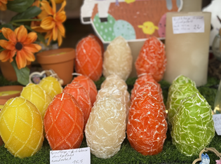 Colorful, textured candles shaped like eggs are displayed in rows on a grassy surface. Background shows decorative pots and vibrant orange flowers.