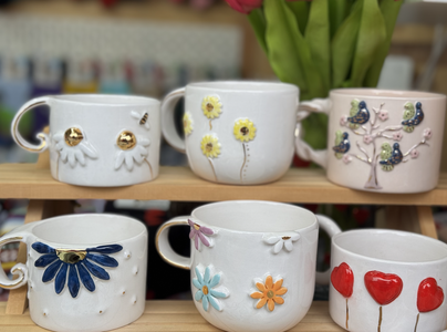 Five ceramic mugs with embossed floral and animal designs are displayed on wooden shelves in a vibrant, cheerful arrangement.