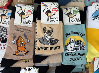 Colorful novelty socks with playful designs featuring a schnitzel, a historical figure, and a cat. Labels read "Socky Wocky" with a jumping monkey.