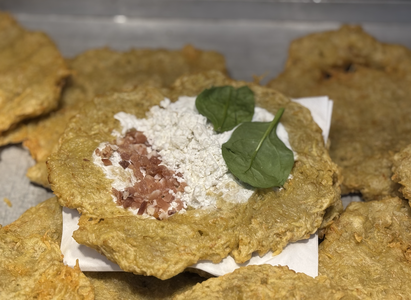Close-up of a golden-brown potato pancake topped with cream, chopped bacon, and spinach leaves, placed on a white napkin.