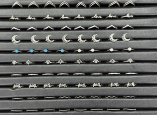 Rows of earrings featuring words "love" and "faith," crescent moons, blue stones, small beads, and flower designs on a black display.