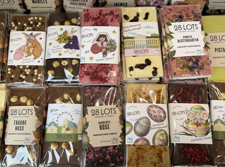 A colorful assortment of 28 LOTS chocolate bars. Varied toppings like nuts, berries, and flowers. Labels feature whimsical illustrations and vibrant designs, creating a playful and enticing display.