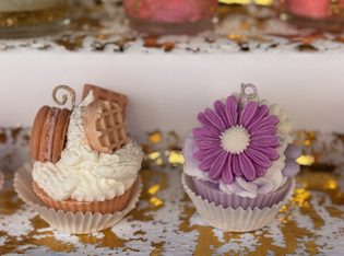 Two decorated cupcakes: one with macarons and a waffle, the other with a purple flower. Both have detailed icing and are on a white shelf.