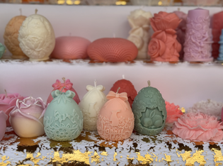 A variety of ornate, pastel-colored candles shaped like eggs and roses, displayed on white shelves with gold leaf accents.