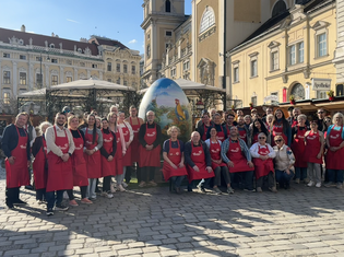 A group of people wearing red aprons pose in front of a large, decorated egg in a European square with historic buildings.