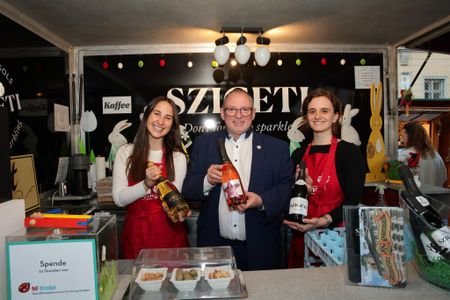 Three people smiling at a booth, holding bottles of sparkling wine. The booth is decorated and has a sign reading "SZIGETI."