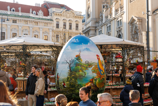 People explore an outdoor market with a large painted egg centerpiece and historic buildings in the background.