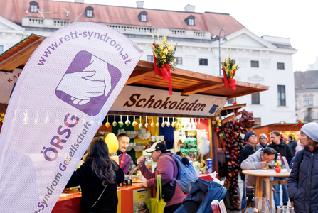 People gathered at a chocolate stall in an outdoor market, with a banner for ÖRSG Rett Syndrome Society in the foreground.