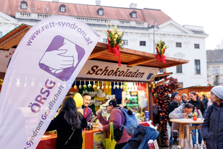 People gathered at a chocolate stall in an outdoor market, with a banner for ÖRSG Rett Syndrome Society in the foreground.