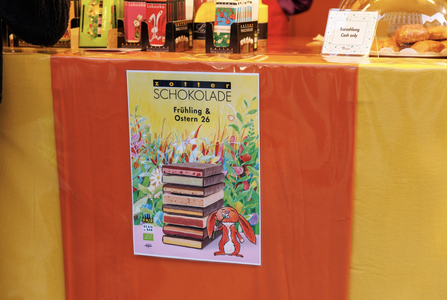 Poster of stacked chocolate bars with colorful flowers and an Easter bunny backdrop on a table at a chocolate shop display.