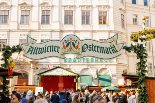 Outdoor market with a decorative sign reading "Alt Wiener Ostermarkt," surrounded by a crowd, historic buildings, and festive decorations.