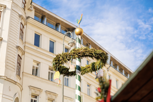 A festive pole decorated with a green wreath and ornaments stands against a clear blue sky, with a historic building in the background.