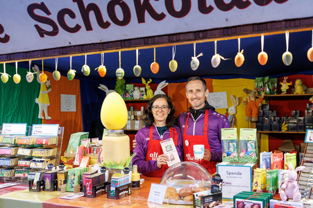 Two people in aprons stand at a colorful chocolate stall, surrounded by Easter decorations and chocolate products.