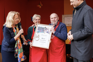 Four people stand together, two holding a framed award. A woman speaks into a microphone. They're on a stage with a red background.