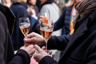 Two people holding glasses of rosé wine amidst a crowded social gathering, wearing dark winter clothing.