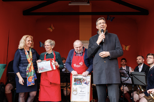 A group of people at an event. A man speaks into a microphone, while others hold certificates. Musicians are seated in the background.
