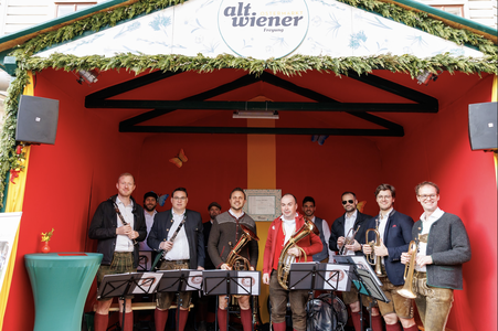 A group of musicians in traditional attire stand with instruments on a stage with red walls and a sign that reads "alt.wiener."
