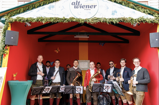 A group of musicians in traditional attire stand with instruments on a stage with red walls and a sign that reads "alt.wiener."