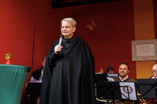 A person in a black robe speaks into a microphone on a stage, with musicians and a red backdrop in the background.