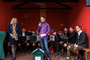 A man speaks into a microphone on a stage with musicians seated behind him, while a woman stands nearby. The setting is decorated with butterflies.