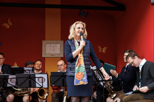 A woman stands on stage speaking into a microphone, with a colorful scarf, in front of a seated brass band. Background has butterflies and red-orange decor.